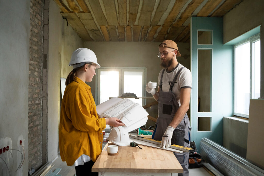 Two construction workers with plans on a table in a room under renovation.