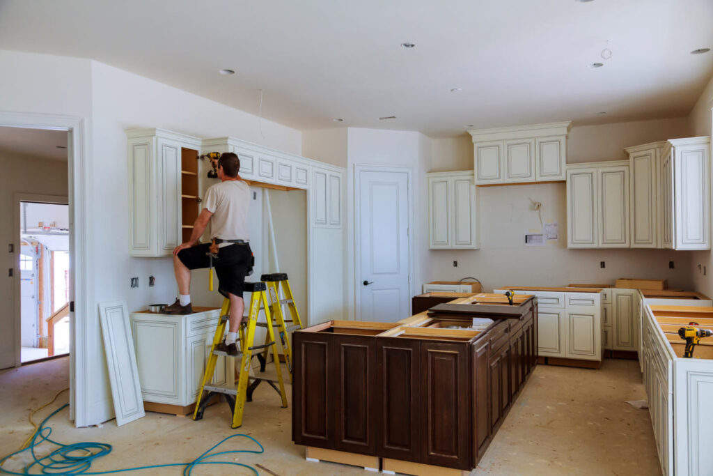 Person on a stepladder working on kitchen cabinets in a room under renovation.