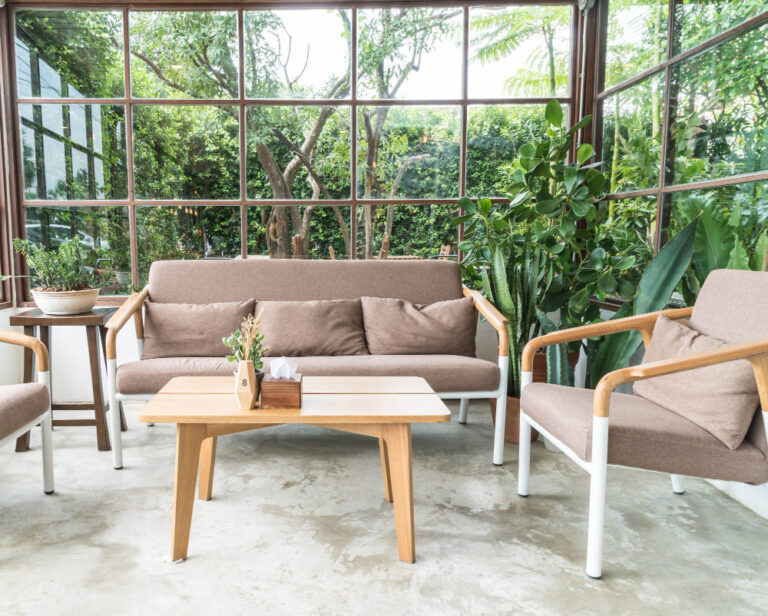 A cozy patio setup with a brown sofa, two chairs, and a center table, surrounded by greenery and large windows.