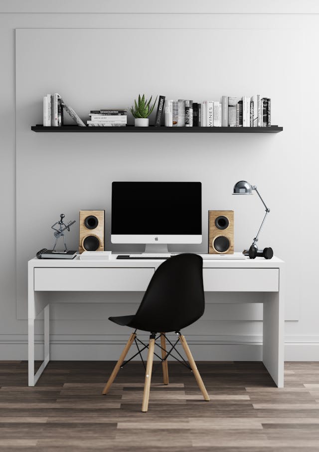 Modern minimalist home office with a white desk, computer, black chair, wooden floor, and wall shelf with books and decor.