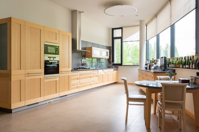 Modern kitchen with wooden cabinets, appliances, and a dining table near large windows.